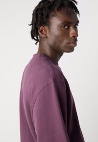 Sudadera púrpura con cuello redondo, hecha de material suave. Presenta un corte relajado y costuras visibles en las costuras de los hombros.