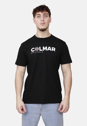 Giovane con capelli corti e ricci che indossa una T-shirt nera Colmar Originals e jeans azzurri, in piedi davanti a uno sfondo chiaro e uniforme.