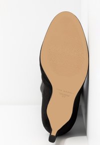 Bottine en suède noire avec un bout pointu et un talon lisse. La semelle est beige, présentant une adhérence à motif et un logo en relief.