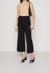 Haut sans manches beige, pantalon noir à jambes larges, sac à main noir texturé, et sandales à talons noirs à talons épais.