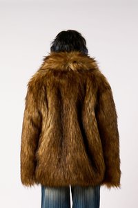 Άτομο με κοντά σκούρα μαλλιά που φορά ένα καφέ ψεύτικο γούνινο παλτό και ξεθωριασμένα μπλε τζιν, γυρισμένο μακριά σε έναν απλό φόντο.