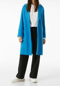 Cappotto lungo blu con cappuccio, caratterizzato da un design strutturato e apertura frontale. Abbinato a pantaloni neri a righe e sneakers bianche.