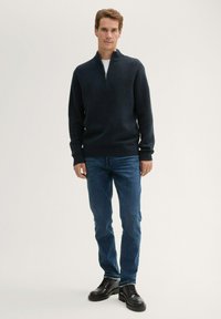 Pull à col zippé marine foncé en tissu texturé, associé à un jean bleu et des chaussures à lacets noires. L'ensemble est décontracté et ajusté.
