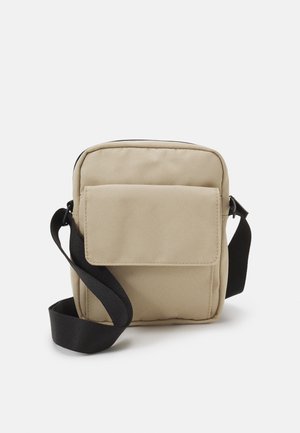 Sac en toile beige avec un dessus à fermeture éclair, poche avant à rabat et sangle réglable noire. Forme rectangulaire avec une texture lisse.