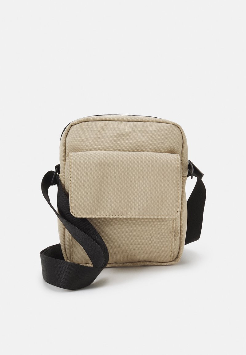 Borsa in tela beige con zip nella parte superiore, tasca a pattina frontale e tracolla nera regolabile. Forma rettangolare con una superficie liscia.