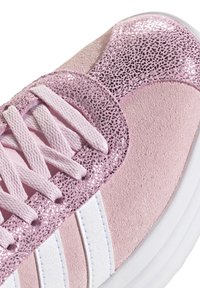 Zapatilla rosa con material de ante, acentos brillantes, rayas blancas y detalles texturizados. Presenta cordones planos en un rosa a juego.