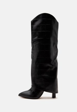 Botte haute en cuir noir avec un motif subtil de crocodile, bout pointu et talon bloc incliné sur fond blanc.