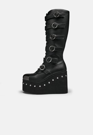 Botte plateforme noire montante jusqu'au genou avec six brides bouclées argentées, laçage à l'avant et semelle compensée cloutée sur fond blanc.