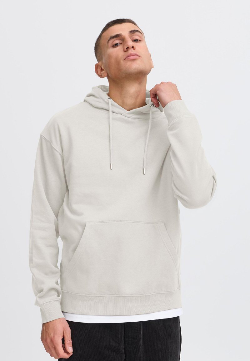 Mladi moški v svetlo bež kapucarju z žepom spredaj, ki si prilagaja vrvico na kapuci, stoji pred enobarvnim belim ozadjem.