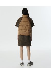 Bruine gewatteerde vest met een hoge kraag, gecombineerd met een donkere korte jurk met een gestructureerd patroon. Gdraagt met grijze sport sneakers.