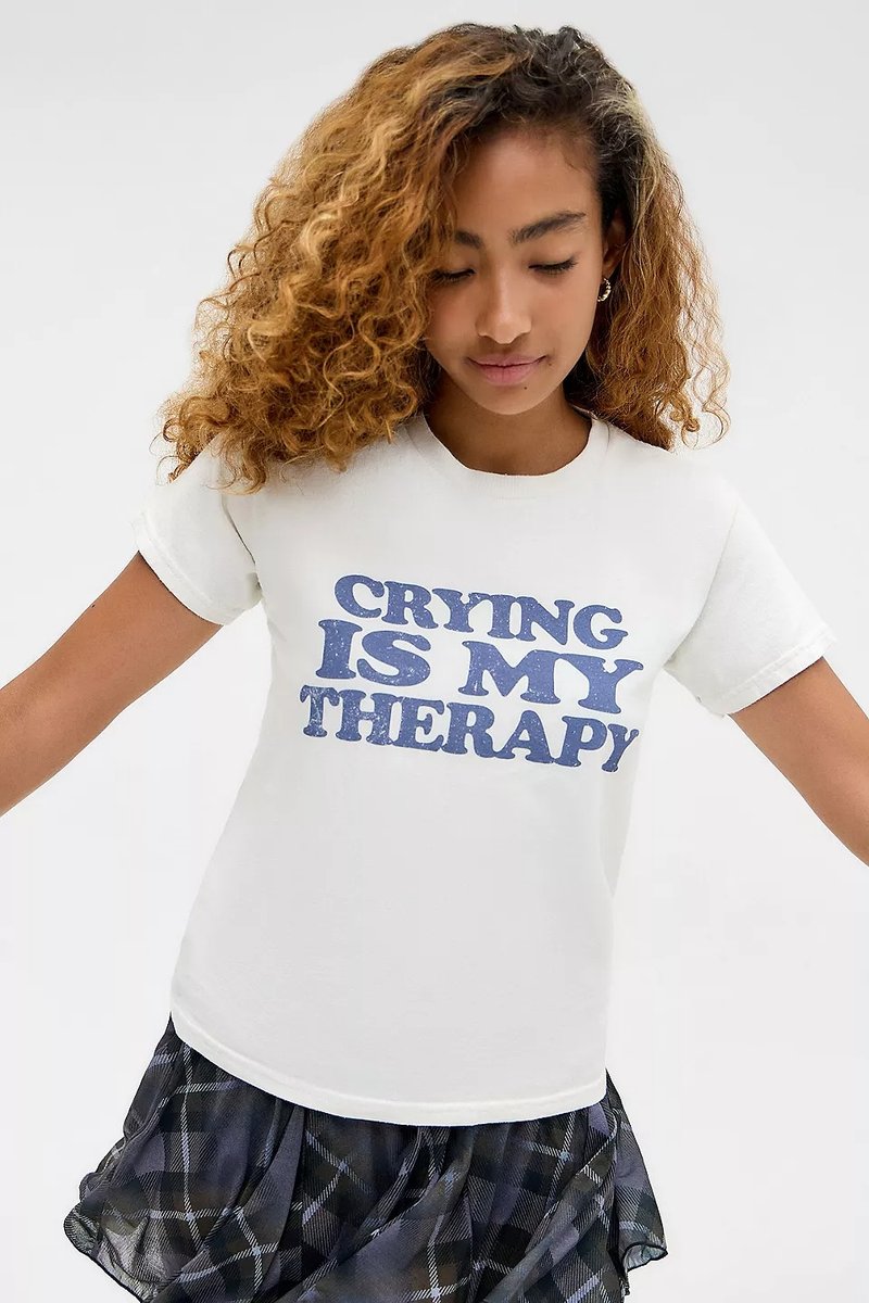 Νεαρή γυναίκα με σγουρά μαλλιά που κοιτάζει προς τα κάτω, φορώντας άσπρο μπλουζάκι με το κείμενο "Το Κλάμα Είναι Η Θεραπεία Μου" και φούστα με καρό σχέδιο.