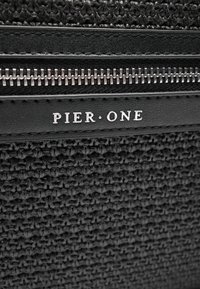Sac en tissu tissé noir avec des accents en cuir lisse, doté d'une fermeture éclair métallique et du logo "PIER ONE" cousu sur une étiquette en cuir.