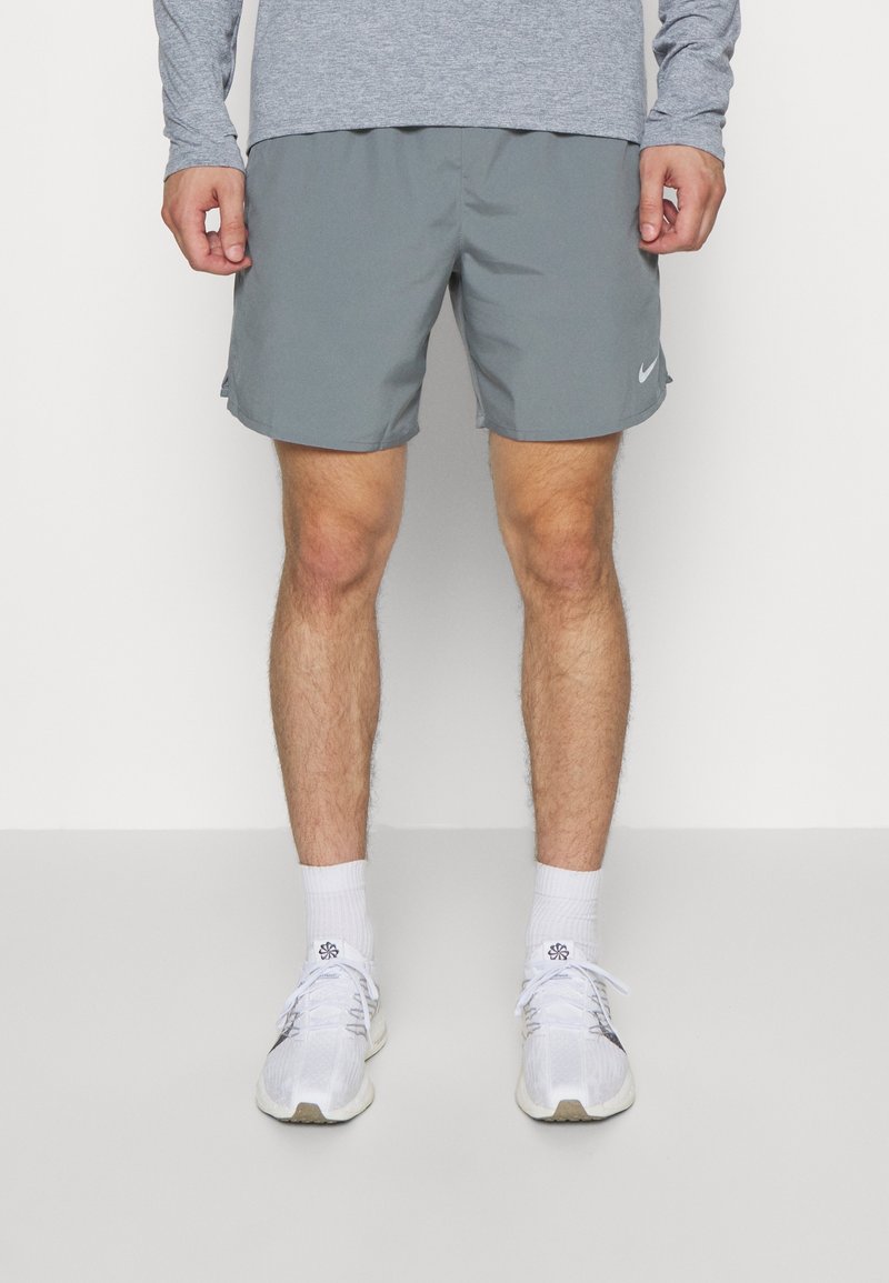 Homme portant un short de sport gris, des chaussettes blanches et des chaussures de course blanches, debout devant un fond clair uni.