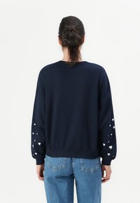 Kvinne med mørkt hår i en bun som har på seg en marineblå genser med hvite hjerte mønstre på ermene og blå jeans, snudd bort fra kameraet mot en hvit bakgrunn.