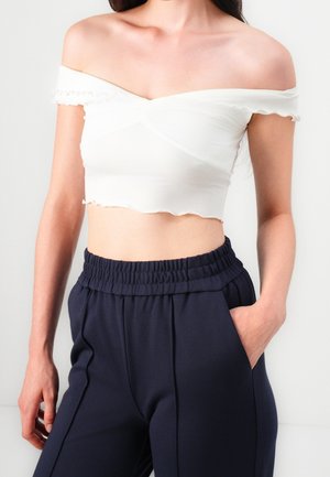 Femme portant un crop top blanc épaules dénudées et un pantalon bleu marine taille haute avec ceinture élastique, une main dans la poche.