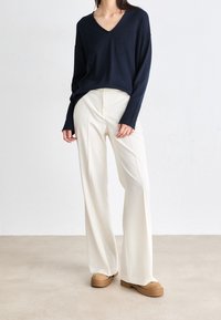 Kobieta w luźnym, granatowym swetrze z dekoltem w serek, białych szerokich spodniach oraz beżowych butach na platformie, stojąca na beżowej podłodze przed białą ścianą.