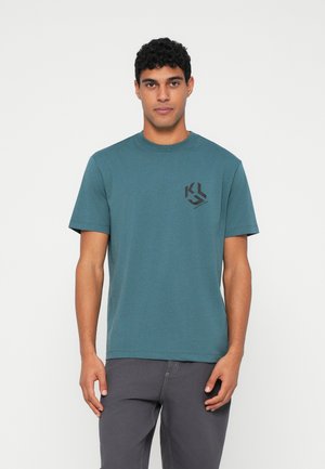 Teal bomulds t-shirt med korte ærmer, rund halsudskæring og et sort logo på venstre bryst. Modellen er stylet med grå bukser. Minimalistisk design.