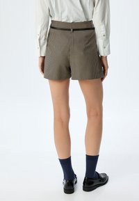 Pantaloni corti a quadri marroni e beige con vita alta, tasche laterali e una cintura nera, abbinati a calzini blu navy e scarpe nere.
