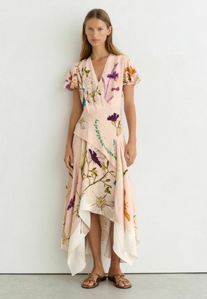 Femme portant une robe portefeuille fleurie rose clair avec des manches volantées et un ourlet asymétrique, debout devant un mur blanc uni.