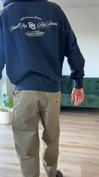 Sudadera azul marino con tipografía blanca en la espalda, combinada con pantalones caqui que incluyen un bolsillo de parche y un acento de logo. Fondo de suelo de madera.