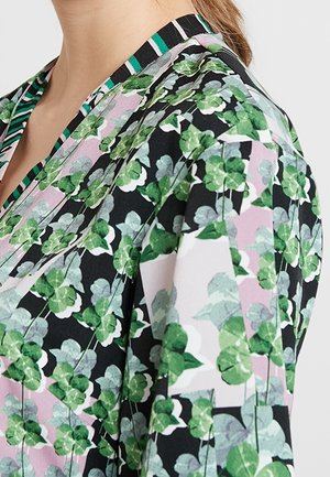Frau trägt eine Bluse mit floralem Muster mit grünen Blättern auf schwarzem, rosa und weißem Hintergrund, zeigt Hals- und oberen Schulterbereich.