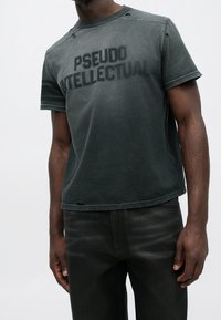Homme portant un t-shirt noir délavé avec les mots "PSEUDO INTELLECTUAL" imprimés sur le devant et des détails usés près du col et de l'ourlet.