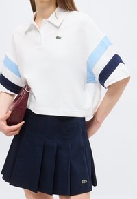 Femme portant un polo blanc court à rayures bleues et une jupe plissée bleu marine, tenant une pochette texturée bordeaux.