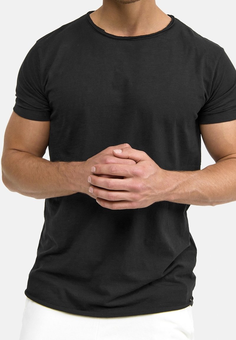 Homme en t-shirt noir à manches courtes et pantalon blanc, debout, les doigts entrelacés à la taille, devant un fond uni.