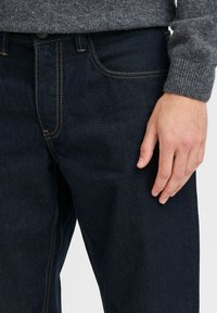 Jeans in denim blu scuro con cuciture arancioni, taglio dritto e design a cinque tasche. Il tessuto ha una texture liscia.