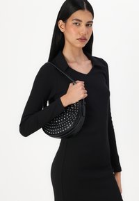 Μαύρη δερμάτινη τσάντα χιαστί σε κυκλικό σχήμα, με καρφιά και ρυθμιζόμενο λουράκι, που κρατάει ένα μοντέλο με φόρεμα μαύρου χρώματος.