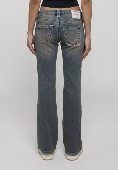 Femme portant un jean bootcut bleu délavé avec des poches arrière cousues et un emblème ailé, vue de dos sur un fond uni.