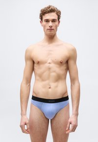 Νεαρός άνδρας με σγουρά μαλλιά που φοράει ανοιχτό μπλε εσώρουχα με μαύρη ζώνη, στέκεται μπροστά σε απλό ανοιχτό γκρι φόντο, κοιτάζοντας μπροστά.