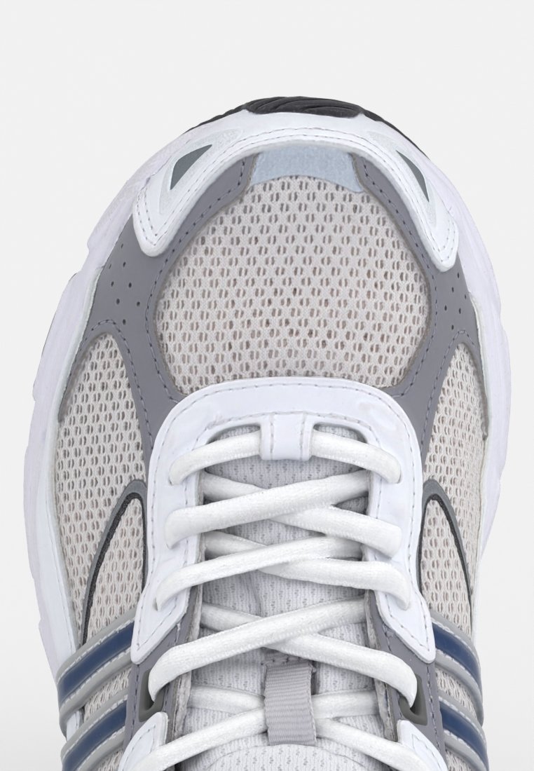 Zapatilla deportiva de malla blanca con acentos grises, que cuenta con una puntera reforzada, cierre con cordones en la parte delantera y múltiples agujeros de ventilación en la parte superior de malla.