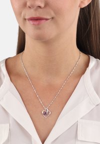 Collana a forma di cuore in argento con una pietra preziosa viola al centro, caratterizzata da un design a catena attorcigliata. Indossata sopra una camicia bianca con colletto.