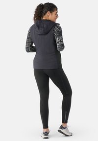 Black padded vest with hood, paired with patterned long sleeves and black leggings. Features zippered ankle openings and athletic sneakers.