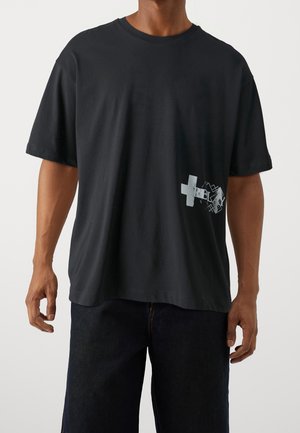 Homme portant un t-shirt noir ample avec une petite croix blanche et un design abstrait près de la taille du côté droit, associé à un pantalon foncé.