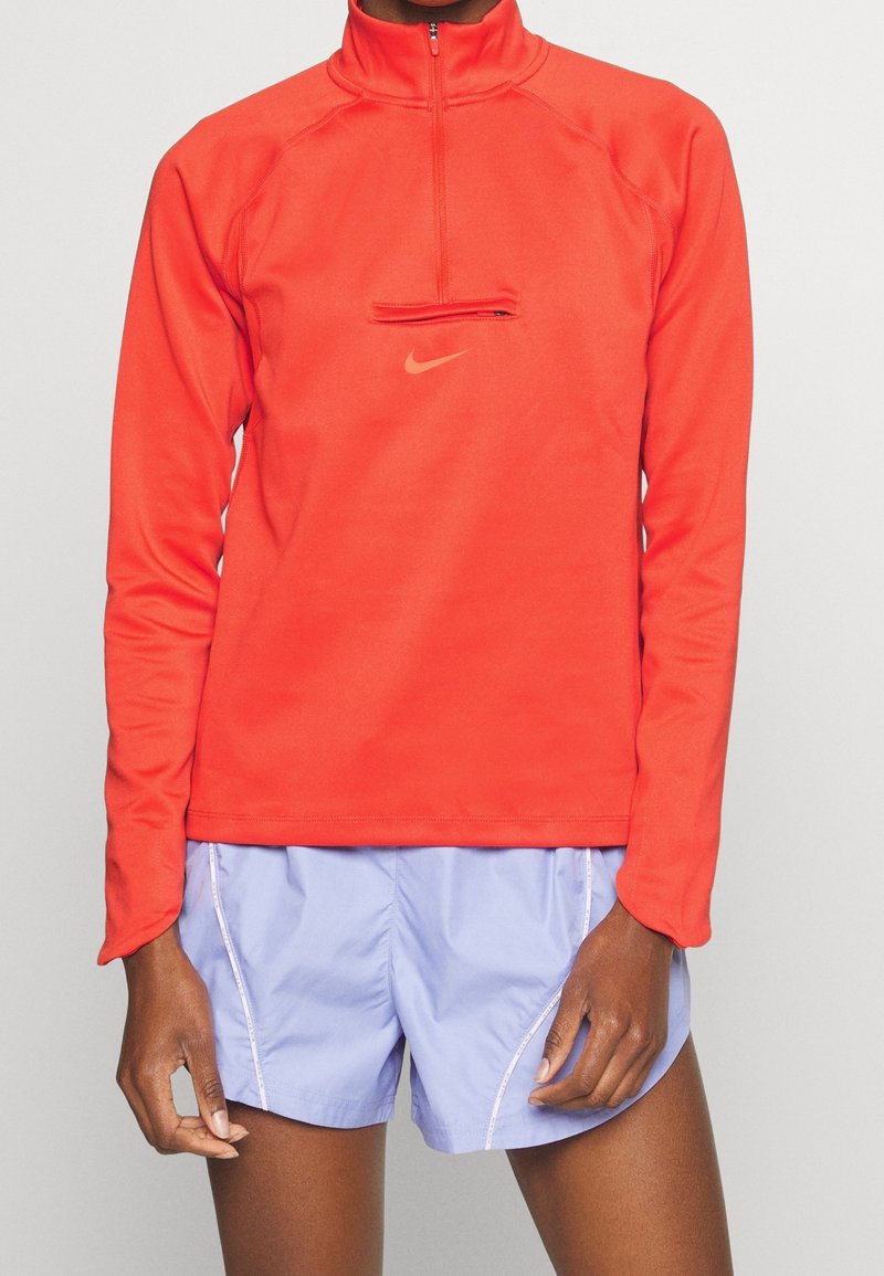 Rotes Langarm-Shirt mit Reißverschluss aus glattem Stoff, mit kleiner Brusttasche und Nike-Logo. Kombiniert mit hellblauen Sportsch shorts.