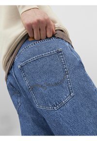 Světle modré džíny z denimu s mírně vybledlou texturou, které mají jedno zadní kapsu s žlutým prošíváním a hnědou etiketou na pase.