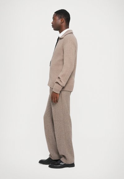 Pull en beige avec un col large et en maille, associé à un pantalon ample en tissu texturé marron clair et des chaussures noires, présenté de profil.