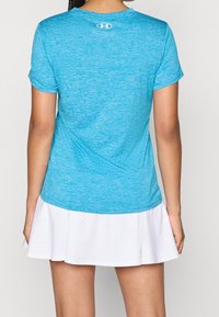 Nainen, joka pukeutuu harmaansiniseen Under Armourin lyhythihaiseen paitaan ja valkoiseen rei’itettyyn tennishameeseen, kuvattuna takaa.