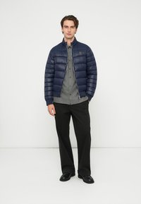 Tmavomodrá páperová bunda s horizontálnym prešívaním, vysokým golierom a zapínaním na zips cez sivú košeľu na gombíky, v kombinácii s čiernymi nohavicami a topánkami.