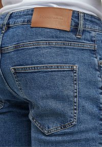 Közeli felvétel kék farmernadrág hátsó zsebének, barna bőr címkével, amelyen az áll: "2Y Premium", fehér inggel viselve.