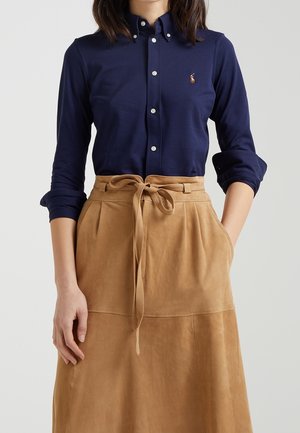 Femme portant une chemise bleu marine à boutons avec les manches retroussées et une jupe taille haute beige nouée devant par un nœud.
