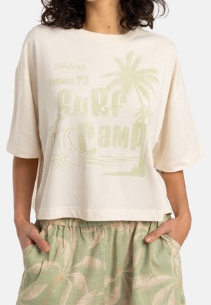 Camiseta holgada color crema en algodón suave, con texto "Surf Camp" y gráfico de palmera estampados en verde. Mangas cortas y dobladillo corto.