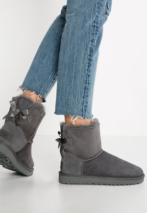 Bottes mi-mollet en daim gris avec doublure en fourrure et nœuds en satin, portées avec un jean bleu court effiloché sur fond blanc.