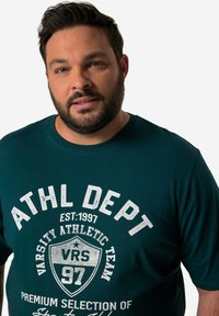 Tealová bavlněná tričko s kulatým výstřihem má bílý grafický potisk a nápisy, včetně detailů "ATHL DEPT" a "EST. 1997".