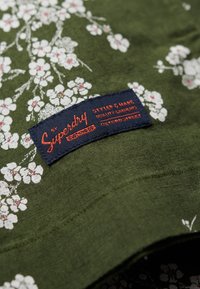 Groene stoffen shirt met witte bloemenpatroon, voorzien van een marineblauwe label met de tekst "Superdry" en aanvullende informatie over kwaliteit en stijl.