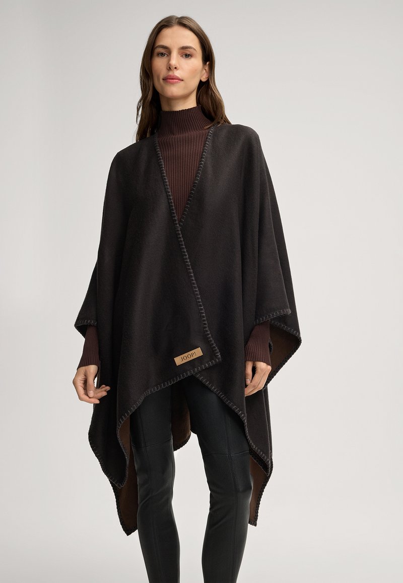 Zwarte poncho gemaakt van zachte stof, met een contrasterende gestikte rand en een klein logo-label. Gekocht over een geribde bruine coltrui.