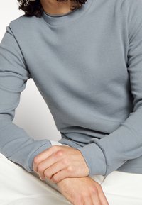 Grauer Waffelstrick-Pullover mit rundem Ausschnitt und langen Ärmeln. Verfügt über gerippte Bündchen und Saum, kombiniert mit hellen Hosen.