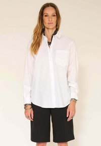 Chemise blanche à boutonnière avec une poche à l'avant, associée à un short noir. La chemise présente un col classique et des manches longues.